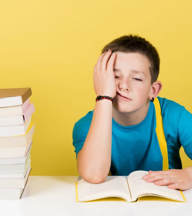 Demotivated boy reading book isolated on yellow background. Bored student holding head with hand next to pile of books